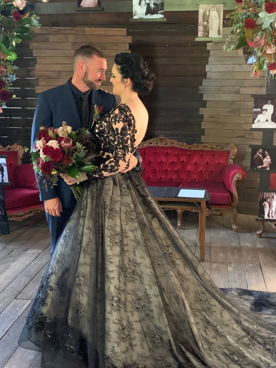 Bride in Maggie Sottero black lace gown with nude underlay and long sleeves embracing her groom in a navy suit at a rustic ceremony venue — bridal alterations by Jacqui