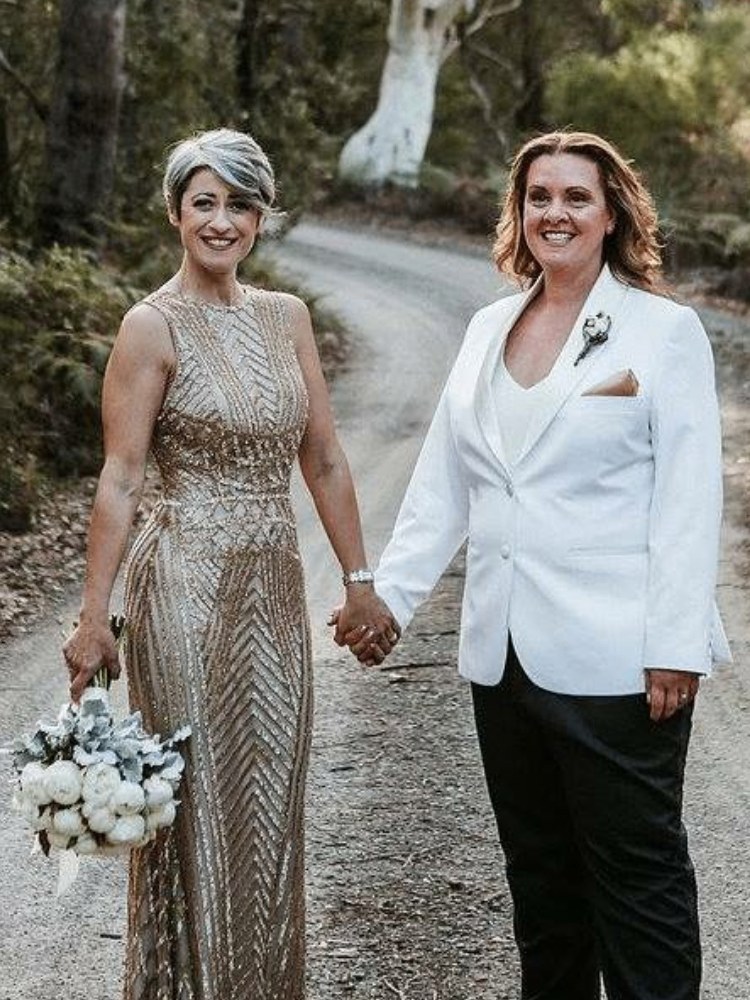 Couple on a bush road — one partner in a floor-length gold beaded gown holding a white bouquet, the other in a white tuxedo jacket and black trousers — formal wear alterations and tailoring by Jacqui