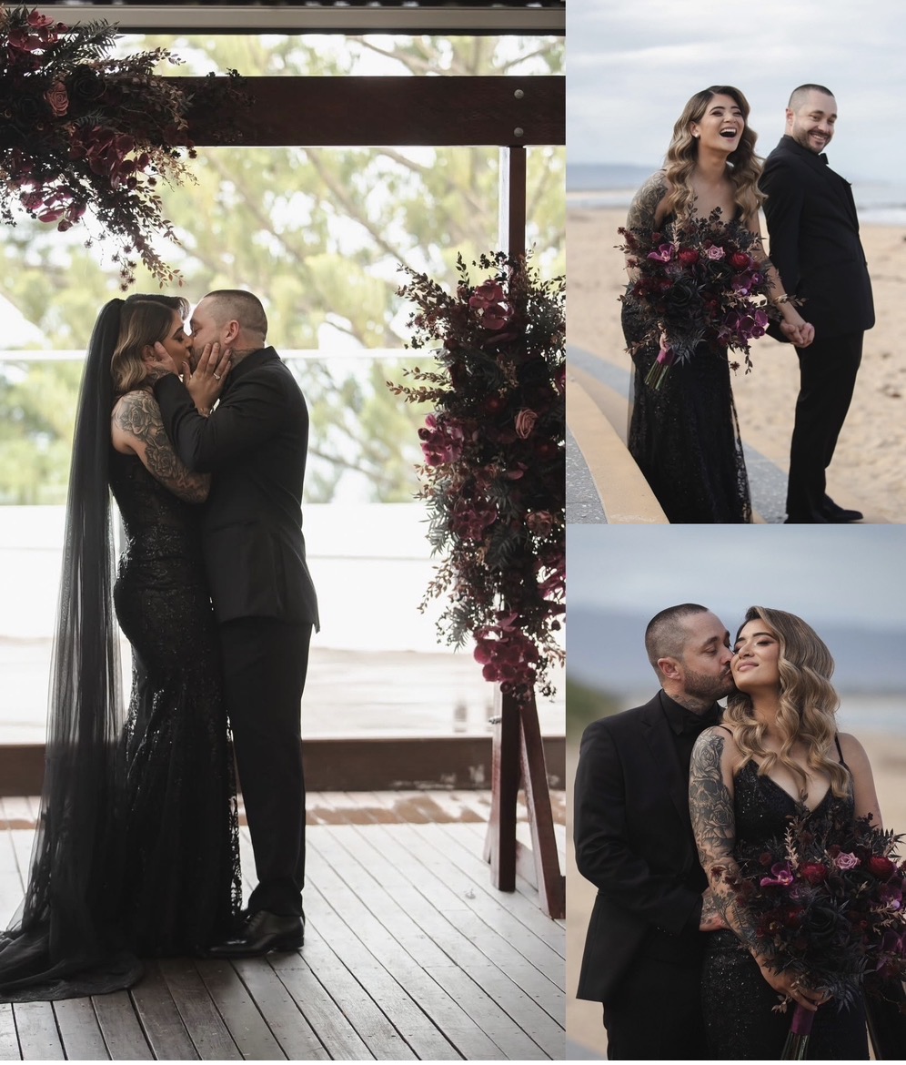 Tattooed bride in a black lace mermaid wedding dress with long black veil kissing her groom in a black tuxedo under a burgundy floral arch — black wedding dress alterations, black veil and bustle by Jacqui