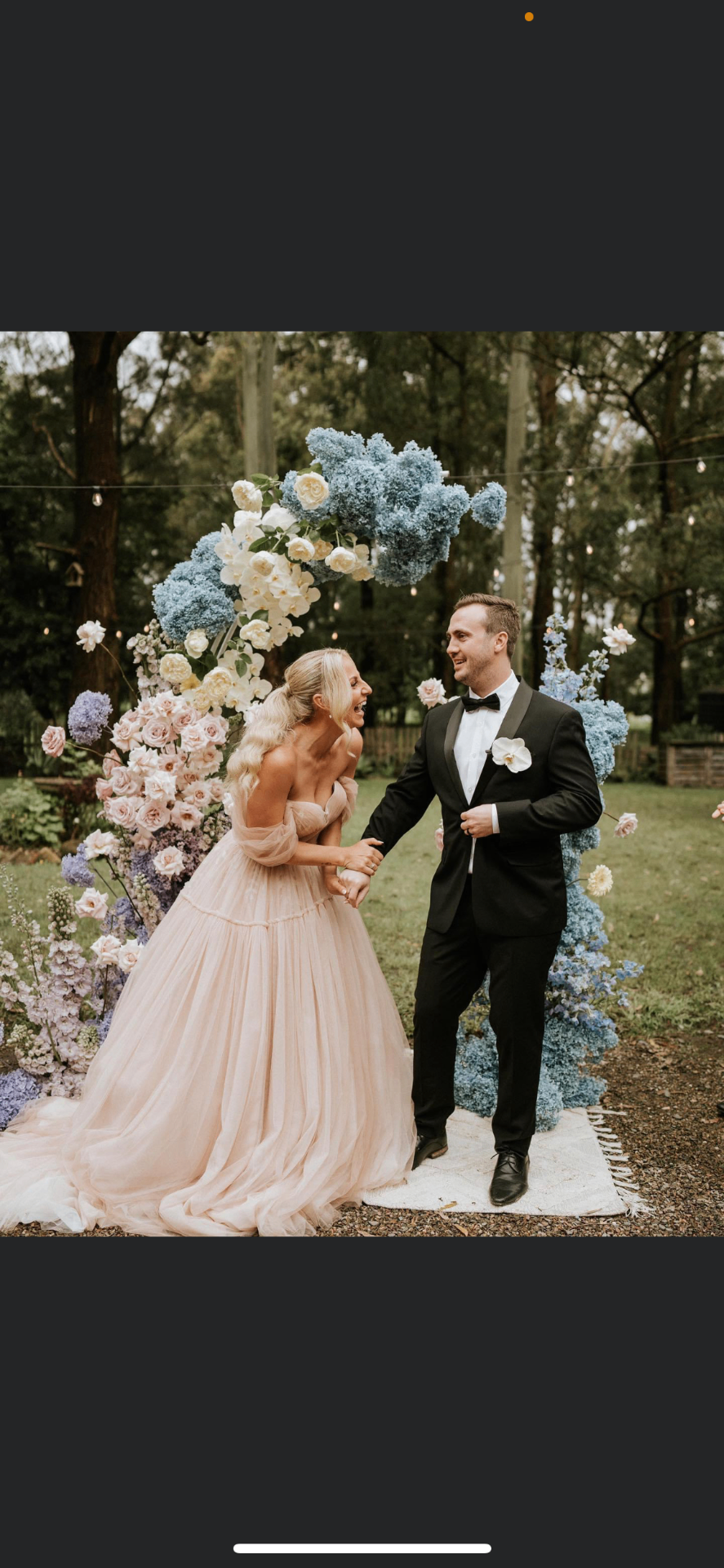 Bride in blush tulle ball gown laughing with groom in black tuxedo under a floral ceremony arch after bridal gown alterations