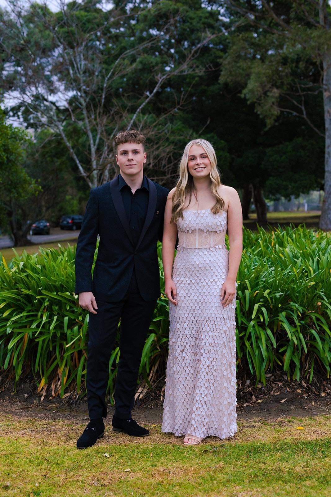 Formal couple — bride in blush scalloped gown and groom in black tuxedo on a garden lawn — formal wear alterations and bridal gown tailoring by Jacqui