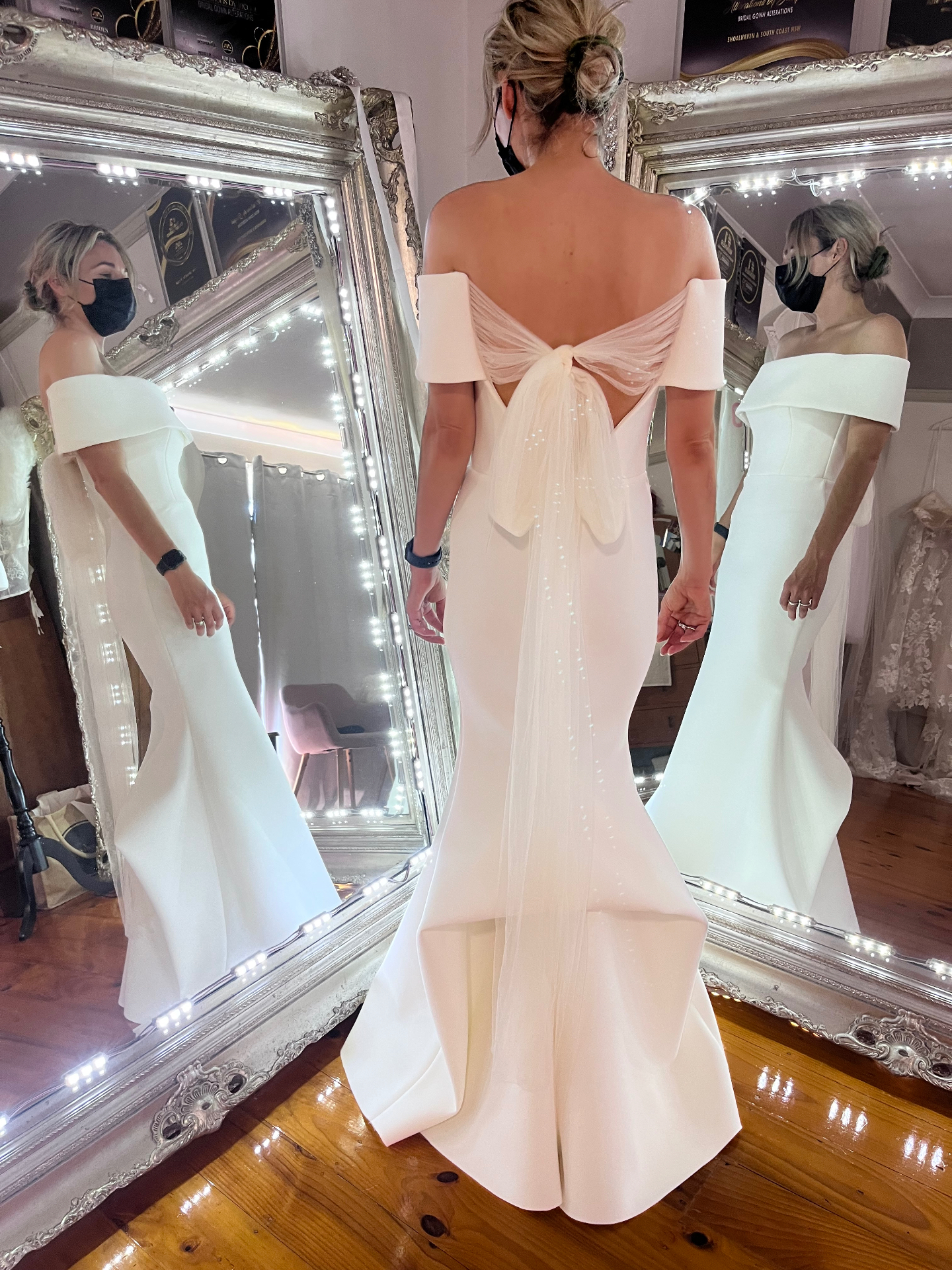 Off-shoulder ivory crepe mermaid wedding gown with sheer tulle bow back detail in a bridal fitting studio — bodice alterations, custom tailoring and unique bustle added for dancing by Jacqui